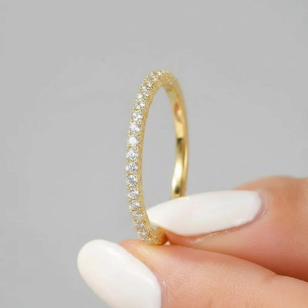 Gold ring with diamonds held by a hand with white nail polish against a blurred floral background